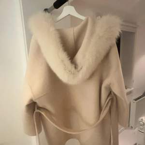 Säljer en elegant beige kappa med en lyxig pälshuva. Den har långa ärmar och ett bälte i midjan för en snygg passform. Perfekt för kyliga dagar när du vill hålla stilen.