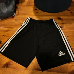Svarta shorts från Adidas - Snygga svarta Adidas shorts med klassiska vita ränder längs sidorna. Perfekta för träning eller en avslappnad dag. Elastisk midja för extra komfort.