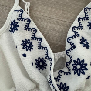 Vit topp med blå broderi - Charmig vit topp med justerbara axelband och vackert blått broderi på bröstet.  