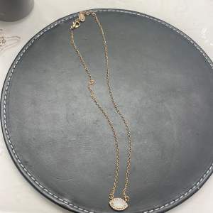 Elegant guldhalsband med en vit oval sten i mitten, omgiven av små glittrande detaljer. Perfekt för att ge en touch av elegans till din outfit.