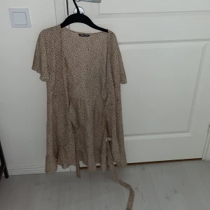Beige prickig klänning - Säljer en beige klänning med prickigt mönster. Klänningen har korta ärmar och ett knytband i midjan för en fin passform. Perfekt för en avslappnad stil.