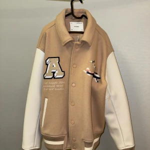 Beige och vit jacka från Axel Arigato | Large - Snygg beige och vit jacka från Axel Arigato med broderade detaljer. Jackan har ett stort 'A' på framsidan och texten 'Arigato' på baksidan. Den är långärmad med knappar och har en cool broderad flygplansdetalj.