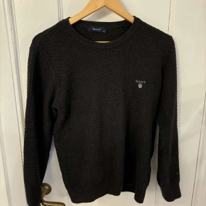 Svart stickad tröja från Gant - Snygg svart stickad tröja från Gant med rund halsringning och diskret logga på bröstet. Perfekt för kyligare dagar och ger en stilren look.