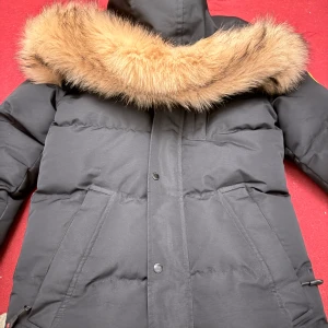 Svart dunjacka med pälsdetalj från Zavetti Canada - Säljer en svart dunjacka från Zavetti Canada med en snygg pälsdetalj på huvan. Jackan har en dragkedja och knappar framtill samt flera praktiska fickor. Perfekt för kalla vinterdagar.