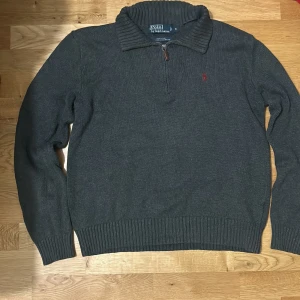 Grå tröja från Polo Ralph Lauren - Säljer en stilren grå tröja från Polo Ralph Lauren i bomull. Tröjan har en dragkedja vid halsen och ribbade detaljer vid krage, ärmslut och nederkant. Perfekt för kyligare dagar. för mer information eller bilder, kontakta mig.
