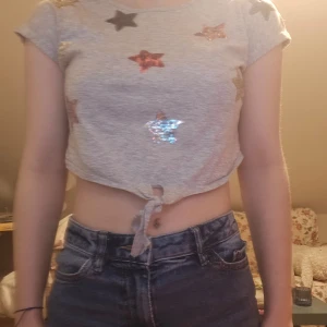 Grå croppad t-shirt med stjärnor - Snygg grå croppad t-shirt med glittriga stjärnor i olika färger. T-shirten har korta ärmar och en knytning framtill som ger en trendig look.