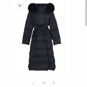 Svart lång dunjacka med pälsdetalj - Elegant svart lång dunjacka med en lyxig pälsdetalj på huvan. Jackan har ett bälte i midjan. Nypris 2300kr.