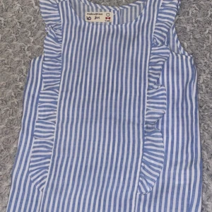 Blå- och vitrandig ärmlös topp - Charmig blå- och vitrandig topp från Made with Love. Den är ärmlös med volangdetaljer längs sidorna och knappar på baksidan. Perfekt för somriga dagar!