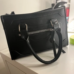 Svart handväska  - Snygg svart handväska i syntetmaterial med praktisk dragkedja framtill. Väskan har två handtag och stilrena detaljer i metall. Perfekt för att bära dina nödvändigheter med stil. 