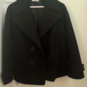 Svart skepparkavaj - Snygg svart skepparkavaj med dubbelknäppning och bred krage. Perfekt för kyligare dagar och ger en stilren look. Passar bra till både jeans och mer formella kläder.