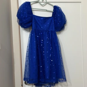 Blå klänning med hjärtmönster - Charmig blå klänning från Monki med puffiga ärmar och hjärtmönster. Klänningen har en knytning i ryggen och är perfekt för en lekfull stil. Den är tillverkad i ett lätt och luftigt material.