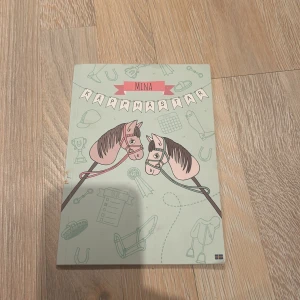 Mina Käpphästar - En charmig bok från Panduro med titeln 'Mina Käpphästar'. Omslaget pryds av illustrationer av käpphästar och hästrelaterade motiv. Boken är i A5-format och innehåller sidor för att planera och dokumentera käpphästaktiviteter. Perfekt för den hästintresserade!