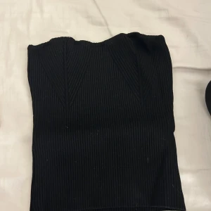 Svart ribbad tubtopp från Gina Tricot - Snygg svart ribbad tubtopp från Gina Tricot. Perfekt för en stilren look och kan enkelt matchas med olika outfits. Toppen är elastisk och bekväm att bära.