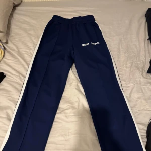 Palm Angels byxor - Snygga blå byxor från Palm Angels med vita sidoremsor och dragkedja vid bensluten. Perfekta för en avslappnad stil. Elastisk midja för extra komfort. Pris kan diskuteras