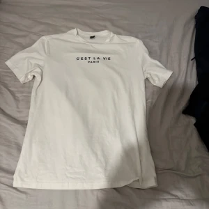 Vit t-shirt med tryck - Säljer en vit t-shirt med texten 'C'est La Vie Paris' tryckt på bröstet. T-shirten har en klassisk rund hals och korta ärmar. Perfekt för en avslappnad stil.