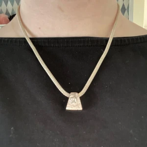 Silverhalsband med triangulär hänge - Elegant silverhalsband med ett triangulärt hänge. Kedjan har en fin flätad design som ger en stilren look. Hänget har en subtil textur som tillför en extra dimension till smycket. Perfekt för att lyfta vilken outfit som helst.