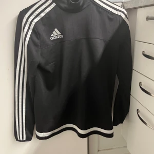 Svart långärmad tröja från Adidas - Säljer en svart långärmad tröja från Adidas med klassiska vita ränder längs ärmarna. Tröjan har en dragkedja vid halsen och är perfekt för sportiga aktiviteter. Adidas-loggan är tryckt på bröstet. Passar ungdomar i storlek L.