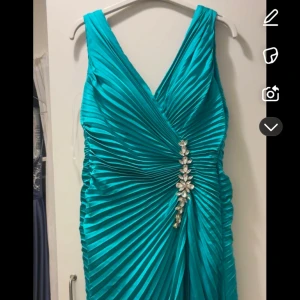 Turkos långklänning med dekorativ detalj - Elegant turkos långklänning med v-ringad design och en glittrande dekorativ detalj framtill. Klänningen har en draperad effekt som ger en lyxig känsla. Original pris 4900