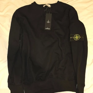 Svart tröja från Stone Island Stl. M - Säljer en helt ny stilren svart tröja från Stone Island med deras ikoniska märke på ärmen. Tröjan har långa ärmar och en klassisk passform. Perfekt för en avslappnad och trendig look.