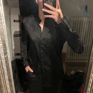 Svart blus/skjorta från H&M - Elegant svart långärmad skjorta från H&M med knappar framtill. Perfekt för en stilren look. Skjortan har en klassisk design med en mjuk och glansig finish.