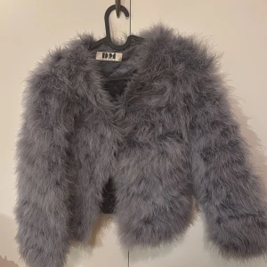 Grå pälsjacka från DennisM - Säljer en fluffig och trendig grå pälsjacka från DennisM. Jackan har en kort och chic design med långa ärmar, perfekt för att ge din outfit en extra edge. Den är idealisk för kyligare dagar när du vill hålla dig varm och stilren.