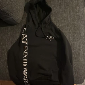 Svart hoodie från Emporio Armani - Snygg svart hoodie från Emporio Armani med logga tryckt i vitt på ärmen och bröstet. Den har en klassisk känguruficka och justerbar huva med dragsko. Perfekt för en stilren och bekväm look. Storlek S men liten i storlek 100%äkta