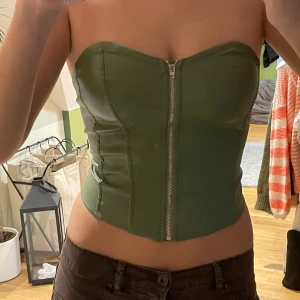 Grön korsett med dragkedja och snörning - Snygg grön korsett med dragkedja framtill och snörning på sidan. Perfekt för en trendig look. Den är axelbandslös och har en tight passform.