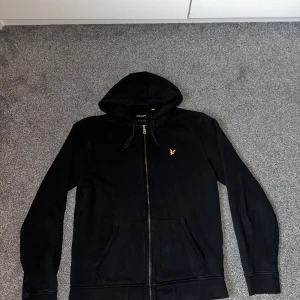 Svart hoodie från Lyle & Scott - Säljer en stilren svart hoodie från Lyle & Scott med dragkedja och huva. Den har ett gult broderat emblem på bröstet och praktiska fickor framtill. Perfekt för en avslappnad look.