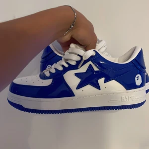 Blå och vita sneakers från BAPE - Snygga blå och vita sneakers från BAPE med en glansig finish. Skorna har en stjärndetalj på sidan och klassisk snörning. Perfekta för att ge din outfit en cool touch. Helt nya ge Bud!