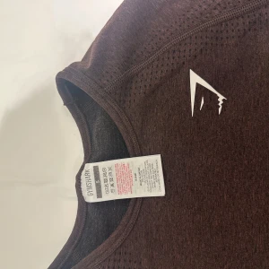 Brun croppad tröja från Gymshark - Säljer en brun croppad tröja från Gymshark. Tröjan har långa ärmar och är tillverkad i ett syntetmaterial som andas, perfekt för träning. Den har en diskret logga på bröstet och en stilren design.