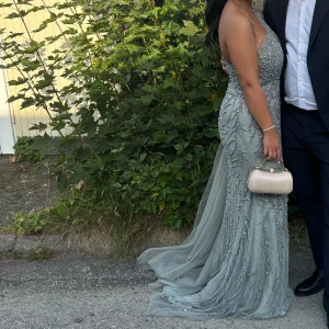 Grå oneshoulder långklänning - Elegant grön långklänning med oneshoulder-design och glittriga detaljer. Klänningen har en slits framtill och är dekorerad med paljetter som ger en lyxig känsla. Samt släp som är avtgbar. Klänningen är i strl 36 och den är strechig. Pris kan diskuteras vid snabb affär.