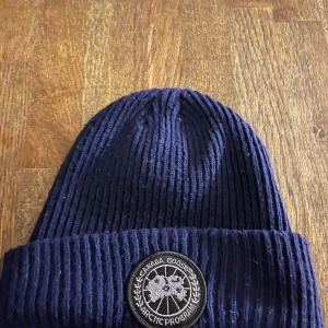 Mörkblå mössa från Canada G*ose - Snygg och varm mörkblå ribbad mössa från Canada Goose med ett broderat märke framtill. Perfekt för kyliga dagar.