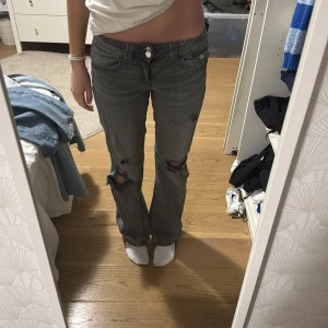 Grå jeans med slitningar - Säljer ett par grå jeans med coola slitningar på knäna. De har en bootcut-stil och är lågmidjade, dem har en liten fläck på framsidan 