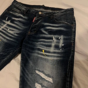 Mörkblå jeans från Dsquared2 - Snygga mörkblå jeans från Dsquared2 med slitna detaljer och färgstänk för en unik look. Klassisk femficksdesign med knappgylf och Dsquared2-logga på bakfickan. Perfekta för en trendig och avslappnad stil. Strl 50