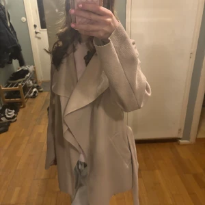 Beige kappa med knytband - Elegant beige kappa med knytband i midjan och långa ärmar. Perfekt för höst och vår, med en stilren design som passar till många tillfällen.