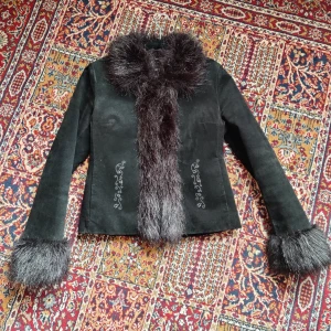 Svart mockajacka - Svartare än svart äkta mockajacka med blombroderier och fuskpälskanter. Stängs med hyskor fram, fickor i sidan finns.  Fodrad med svart satin och nåt fluffigt mellanlager som gör jackan varm och mysig att glida runt i hela hösten ut.  Märkt storlek M