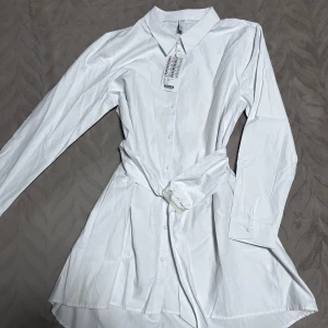 Vit skjortklänning - Säljer en HELT NY stilren vit skjortklänning 💗i storlek XL men det passar på L också. Klänningen har långa ärmar och knappar framtill, samt ett knytskärp i midjan för en snygg passform. 
