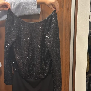 Svart glittrig klänning - Snygg svart klänning med glittriga paljetter och öppen rygg. Perfekt för en festlig kväll. Klänningen har långa ärmar och en elegant drapering baktill. Det är fråm Ados