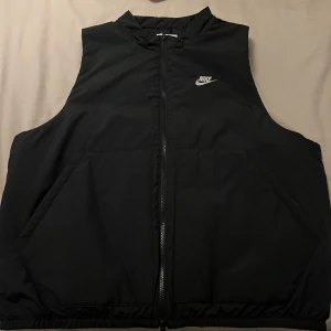 Svart väst från Nike - Säljer en svart Nike Therma-Fit väst med dragkedja och två stora fickor framtill. Perfekt för kyligare dagar och har en stilren design med Nike-logga på bröstet.