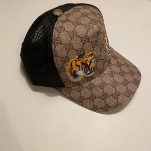 Mönstrad keps med tigertryck - Snygg keps från Gucci med ett djärvt tigertryck på framsidan. Kepsen har ett brunt mönster med märkets logotyp och svart mesh på baksidan för ventilation. Justerbart band i klassiska Gucci-färger för perfekt passform. Skriv ett meddelande för bilder, info etc