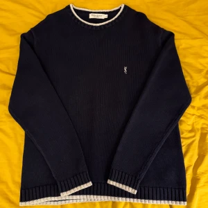 Mörkblå stickad tröja från Yves Saint Laurent - Säljer en stilren mörkblå stickad tröja från Yves Saint Laurent med vita ribbade detaljer vid hals, ärmslut och nederkant.