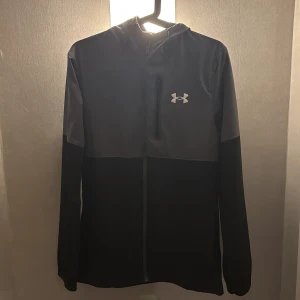Svart och grå jacka från Under Armour - Säljer en stilren svart och grå jacka från Under Armour. Jackan har en dragkedja framtill och en praktisk bröstficka med dragkedja. Jackan är helt oanvänd och 10/10 i skick. Storlek S. Skriv privat vid funderingar!