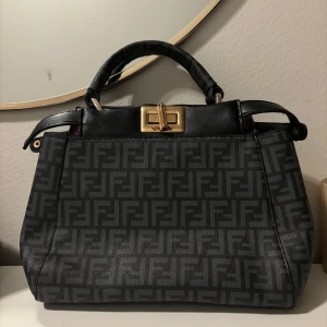 Svart och grå handväska med mönster - Snygg handväska i svart och grått med ett stilrent mönster. Väskan har ett elegant spänne i guld och ett bekvämt handtag. Perfekt för att ge din outfit en lyxig touch.