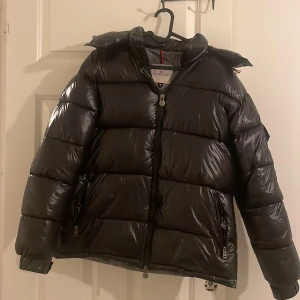 Svart pufferjacka från Moncler - Säljer en snygg svart pufferjacka från Moncler. Jackan har en glansig finish och är utrustad med en huva och dragkedja framtill. Perfekt för kyligare dagar med sin vadderade design.