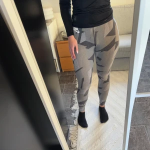 Grå camo leggings från Nike - Snygga grå camo leggings från Nike med ett coolt mönster. Perfekta för träning eller en avslappnad dag. De har en elastisk midja med 'Just Do It' tryckt på insidan. 