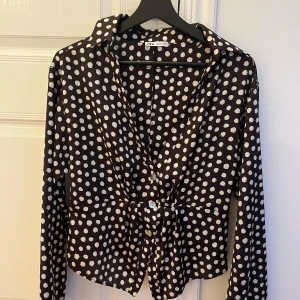 Brun och vit prickig blus från Zara - Snygg svart blus från Zara med vita prickar och långa ärmar. Blusen har en v-ringad design och knappar framtill. Perfekt för en stilren look.