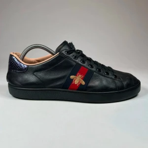 Gucci ace bee - Snygga svarta Gucci ace bee sneakers i skinn med en broderad guldfärgad bi på sidan. Skorna har svarta snören och en röd och blå randdetalj. Perfekta för en stilren look.