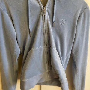 Ljusblå set från Juicy Couture - Säljer ett ljusblå sett från Juicy Couture i mjukt material. Tröjan har dragkedja framtill och en broderad logga på bröstet. Den har också en praktisk ficka och justerbar huva med snören. Perfekt för en avslappnad stil. Byxorna har fickor därback och är i bra skick bara att på ena fickan har en knapp gått av.