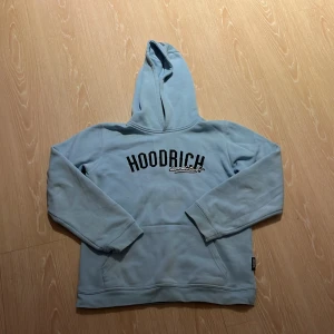 Ljusblå hoodie från Hoodrich - Säljer en snygg ljusblå hoodie från Hoodrich med svart tryck på bröstet. Den har en klassisk design med huva och långa ärmar, perfekt för en avslappnad stil. Passar ungdomar i åldern 13-15 år.