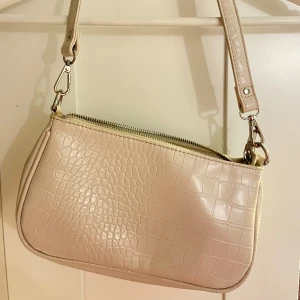 Beige axelväska  - Snygg beige axelväska med krokodilpräglat mönster från Gina Tricot. Väskan har en praktisk dragkedja och en justerbar axelrem. Perfekt för att bära dina nödvändigheter med stil. Lite smutsig inuti, se bilder.